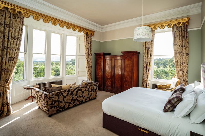 A bedroom with a large double bed with pillow, wooden wardrobe and sofa looking out to a view of trees.