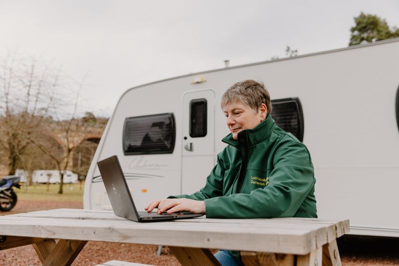 A member of staff on a laptop sitting at picnic table beside a caravan