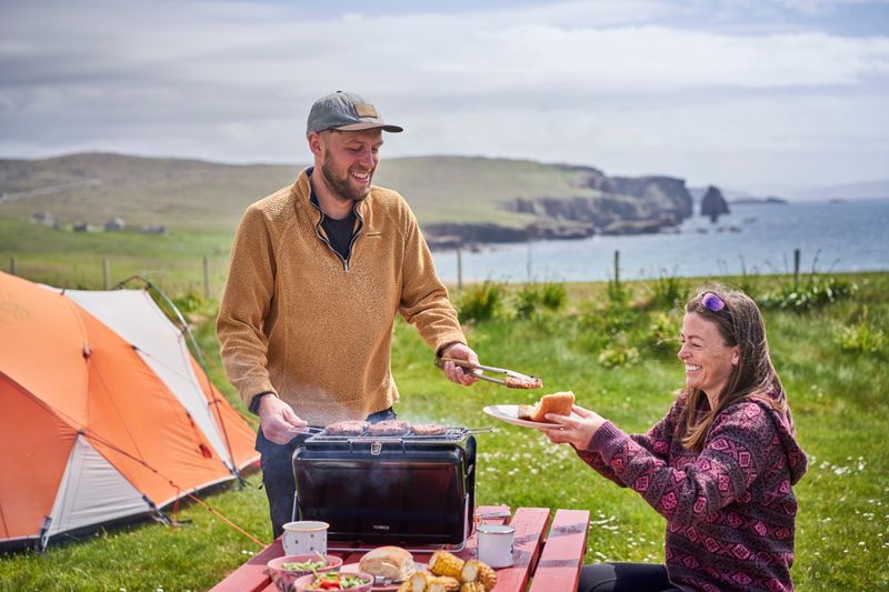 A man and woman sitting at a picnic table, cooking on a barbeque with a tent in background.