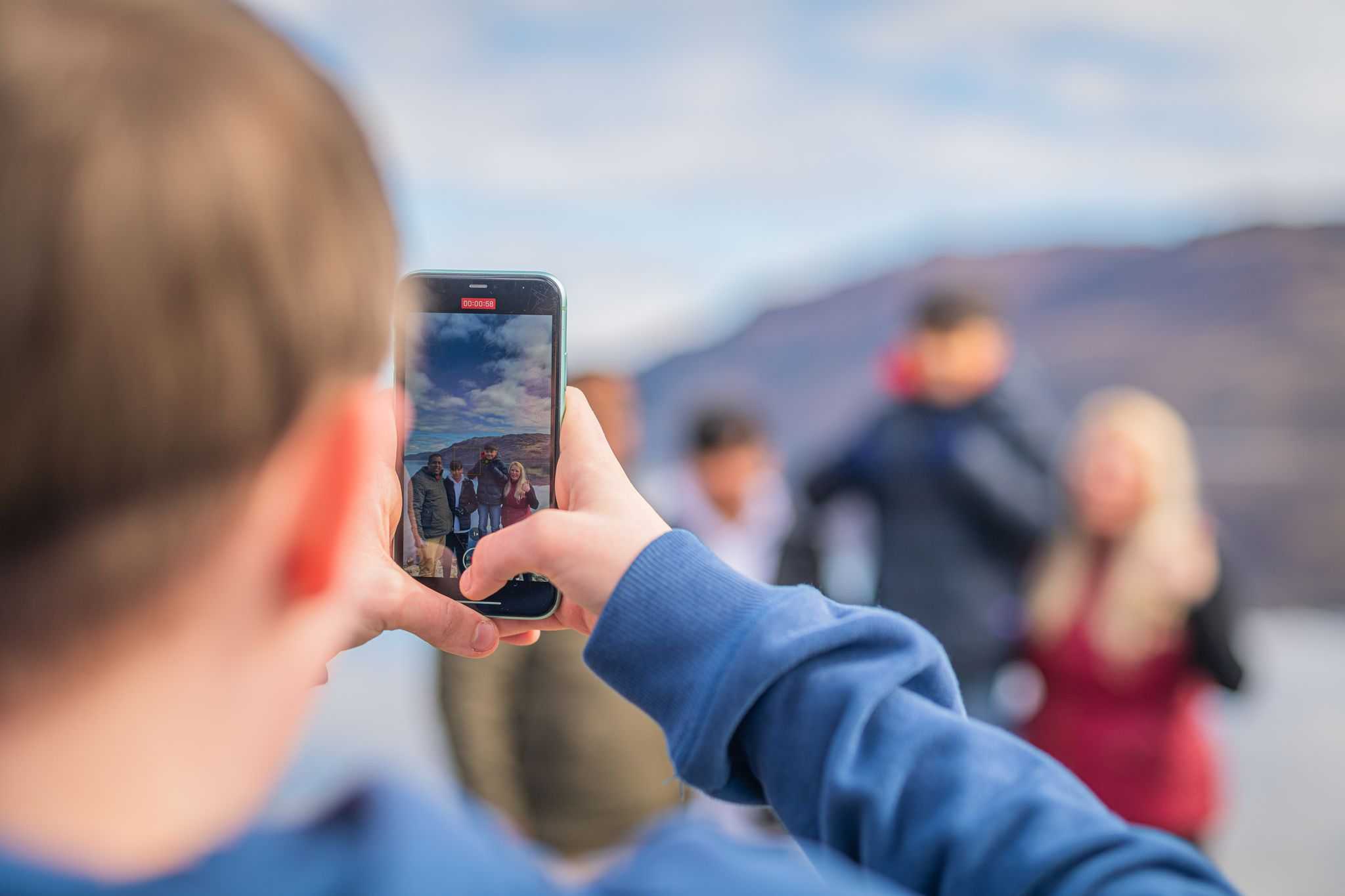 A close up of a man taking a photograph on his mobile of a family stood on a loch pier