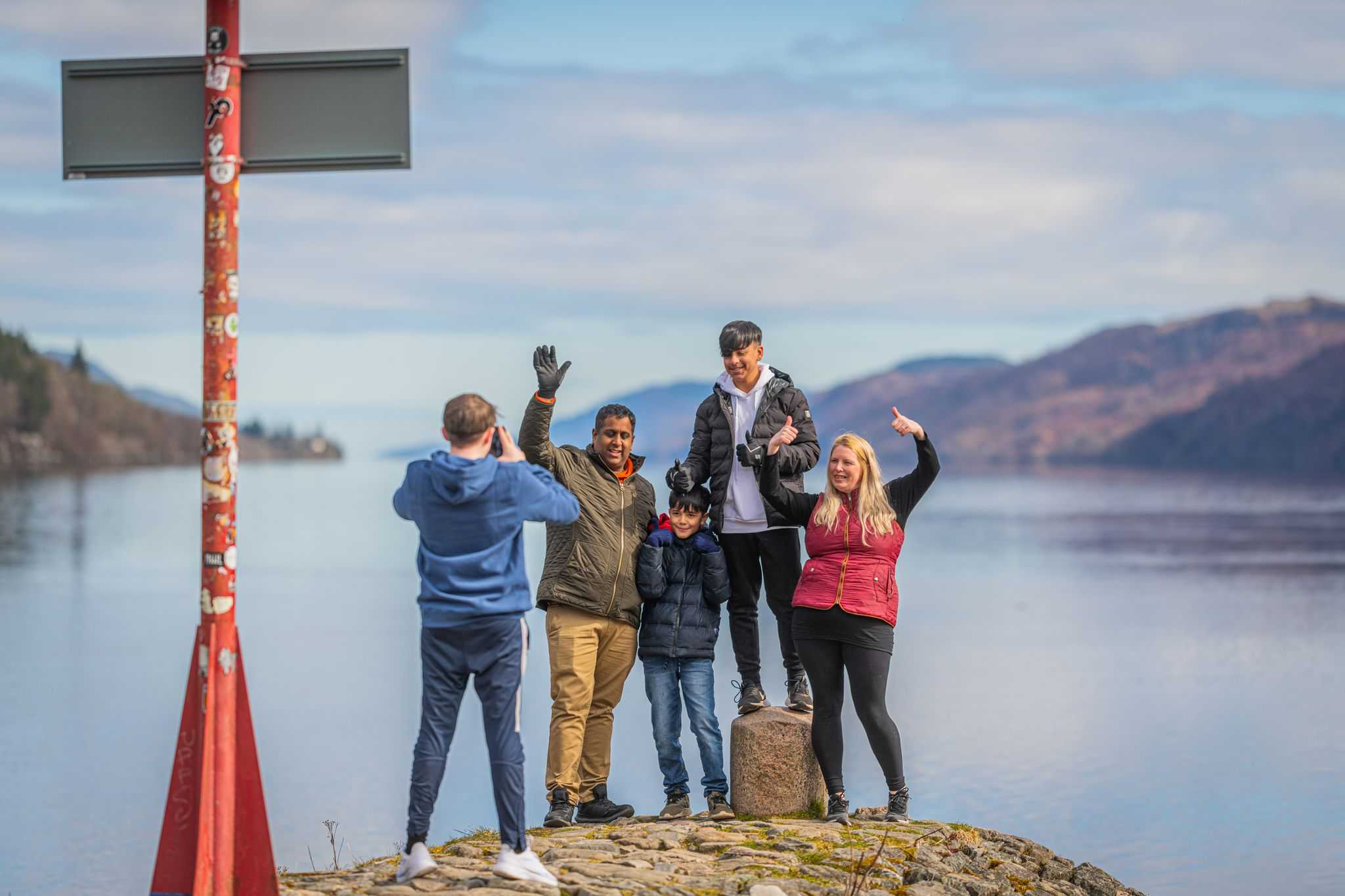 A young man takes a photograph of a family with a mum, dad and two boys on the end of a loch pier